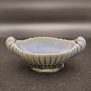 May include: A ceramic, oval-shaped bowl with a light blue interior and a scalloped rim. The exterior is a muted green with vertical ribbed detailing. Two curved handles are on either side. The bowl sits on a small, round base.