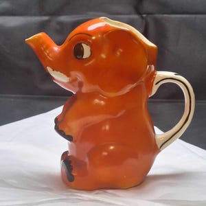 May include: A ceramic orange elephant-shaped pitcher with a white handle and spout. The elephant has a smiling face with brown eyes and black accents on its feet. The pitcher is a decorative item.
