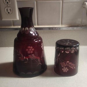 May include: A ruby red glass decanter and tumbler set. The decanter has a bulbous base and a slender neck, with etched floral and grape designs. The tumbler features similar etched patterns. A classic glassware set.