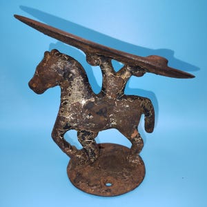 May include: A vintage cast iron boot jack featuring a horse figure with a rider. The metal object has a weathered, dark brown finish with visible signs of rust and chipped paint. It stands on a circular base with a hole for mounting.