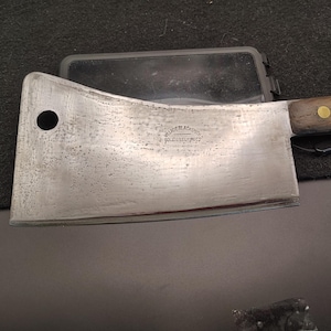 May include: A large, well-worn cleaver with a wooden handle and a metal blade. The blade has a circular hole near the top and the text "VILLAGE BLACKSMITH SOLID STEEL FORGED" is stamped on the blade.