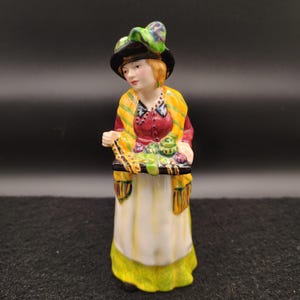 May include: A ceramic figurine of a woman wearing a black hat with a green feather, a yellow and green scarf, and a long white skirt with a green hem. She is holding a tray with green and purple items.