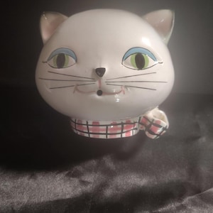 May include: A white ceramic cat-shaped planter or mug featuring painted green eyes with blue eyeshadow, a small black nose, thin black whiskers, and a pink and white checkered scarf pattern around the base. The cat has a rounded head shape.