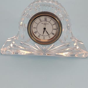 May include: A clear, faceted crystal mantel clock with a rounded arch shape. The clock face is white with black Roman numerals and the text Waterford Crystal. It features a gold-toned metal bezel, black hour and minute hands, and a second hand.