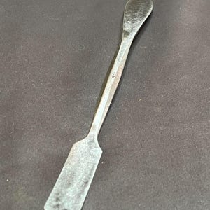 May include: A silver-toned metal spatula with a rectangular blade and a curved handle. The spatula has a diamond-shaped detail on the handle. The tool is designed for spreading or lifting materials.
