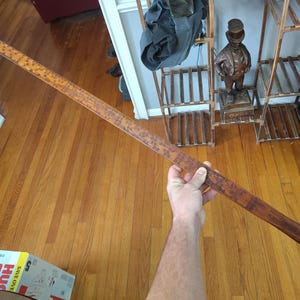 May include: A long, straight wooden yardstick with a dark brown finish and a smooth, rounded end. The surface features clearly printed black numerical markings and measurement increments along its length, showing a scale up to 36 inches.