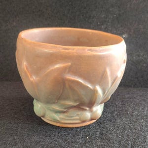 May include: A small, antique ceramic bowl with a matte finish. The bowl is a muted peach color with a green base and leaf-like embossed detailing around the sides. The bowl has a rounded shape and a wide opening.
