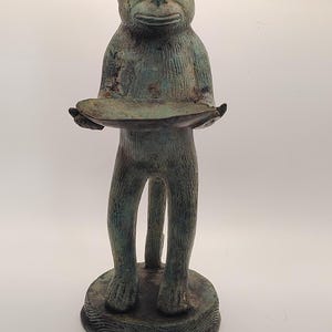 May include: A standing monkey figurine with a weathered, verdigris-toned finish. The monkey is depicted in an upright position, holding a shallow, circular bowl in its hands. It stands on a round, textured base with a subtle wave-like pattern.