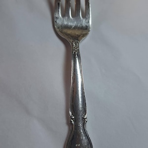 May include: A silver fork with four tines, featuring a decorative handle. The utensil has a slightly tarnished appearance, suggesting age or use. The handle has an ornate floral design.