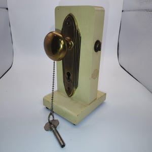 May include: A cream-colored wooden display block featuring a brass door knob and an elongated backplate with a keyhole. A metal ball chain is attached to the knob, holding a silver-toned skeleton key. The side of the block displays a Dexter logo.