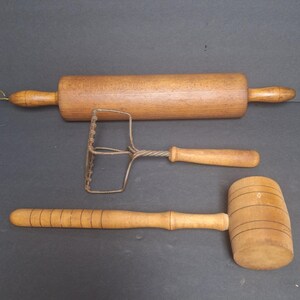 May include: A set of three vintage kitchen tools: a wooden rolling pin, a wire potato masher, and a wooden mallet.