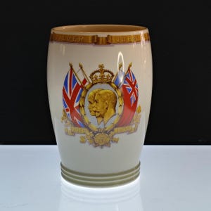 May include: Cream-colored ceramic commemorative mug with a gold rim and base. The mug features a central emblem with profiles of two figures, a crown, and Union Jack flags. The text "SILVER JUBILEE" is displayed across the top, with the years 1910 and 1935.