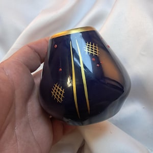 May include: A dark blue, teardrop-shaped ceramic vase with gold trim and accents. The vase features gold vertical lines and crosshatch patterns, along with small red dots. The top rim is also trimmed in gold.