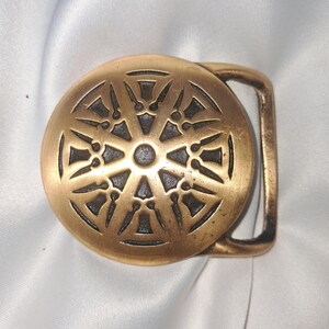 May include: A vintage brass belt buckle with a circular design. The buckle features an intricate pattern of radiating lines and shapes, creating a sunburst or wheel-like motif. The metal has a slightly aged appearance, adding to its vintage appeal.