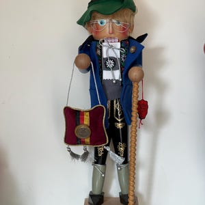 May include: A wooden nutcracker figurine dressed in traditional German clothing. He wears a green hat, blue jacket, black pants, and white socks. He holds a walking stick and a small red bag with a gold coin. The base of the nutcracker reads "1948-2002 Deutsche Gold Mark Last German Coin."
