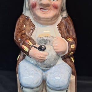 May include: A ceramic figurine of a seated man in a tricorn hat and brown coat. The figure has rosy cheeks and holds a pouch and pipe. The figurine is painted in shades of brown, beige, blue, and white.