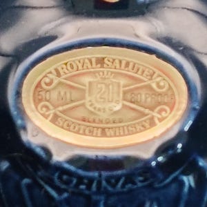 K&ouml;nnte beinhalten: Nahaufnahme einer dunkelblauen Flasche mit goldenem Etikett. Das Etikett lautet "Royal Salute 21 Years Old Blended Scotch Whisky" mit den Ma&szlig;en 50 ml und 80 Proof. Die Flasche hat ein gl&auml;nzendes Finish.