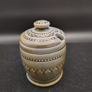 May include: A ceramic sugar bowl with a lid and a spoon slot. The bowl is a muted green-gray color with decorative patterns, including dots, lines, and clover shapes. The lid has a small knob on top.
