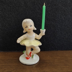 May include: A ceramic figurine of a young girl in a ballet pose, holding a green candle. The girl wears a white top and a yellow tutu. Her ballet shoes are red. The figurine is on a white base.