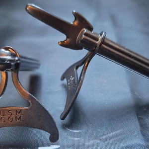 May include: Two silver metal guitar picks with a stand. The stand has the inscription "U.S.M. COM."