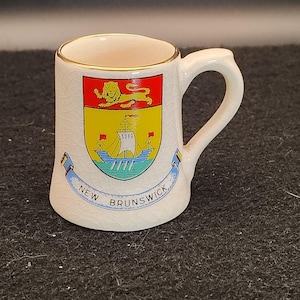 May include: A small, off-white ceramic mug with a gold rim and handle. The mug features a colorful crest with a lion, ship, and the text "NEW BRUNSWICK" on a blue banner. The mug has a slightly cracked, vintage appearance.
