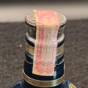 K&ouml;nnte beinhalten: Nahaufnahme einer Vintage-Lik&ouml;rflasche mit silbernem Verschluss und einem rot-beigen Bureau of OPAFE-Siegel. Die Flasche hat einen dunkelblauen K&ouml;rper mit goldenen Akzenten und einem dekorativen Etikett.