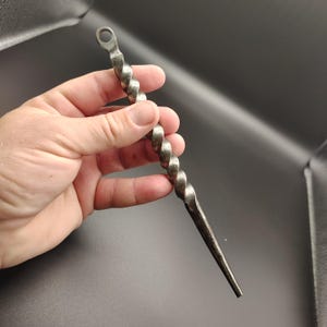 May include: A hand-forged metal bar with a twisted design. The bar has a loop at the top and tapers to a point at the bottom. The metal has a dark, aged finish.
