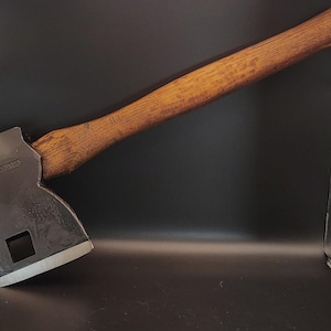 May include: A vintage axe with a wooden handle and a dark metal head. The axe head has a square hole and a sharp blade. The wooden handle is a light brown color and shows signs of wear. The axe is a tool for chopping wood.