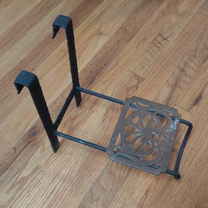 May include: A black metal plant stand with a decorative, openwork design on the base. The stand has two upward-curving hooks and a rectangular base with an ornate floral pattern. The stand is designed to hold a plant pot.