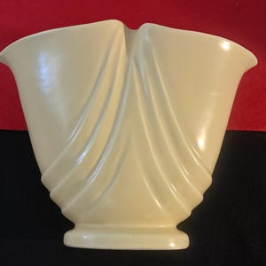 May include: A cream-colored ceramic vase with a unique, draped design. The vase has a flared top and a pedestal base. The front of the vase features vertical, flowing lines that create a draped effect. The vase is a decorative item.