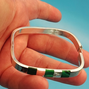 May include: A square-shaped silver hinged bangle bracelet featuring inlaid rectangular segments of dark green malachite stone. The bracelet has a polished metallic finish and a secure side clasp mechanism, creating a geometric design.