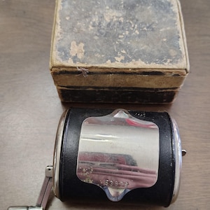 May include: A vintage black and silver pencil sharpener with a crank handle and a decorative silver plate. An antique, rectangular box with a worn, textured exterior sits behind the sharpener.