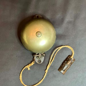 May include: A vintage brass bell with a small loop for hanging. The bell is connected to a decorative wooden tag by a length of natural-colored rope. The tag features a carved design. The bell is a classic decorative item.
