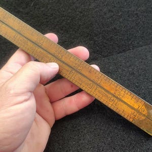 May include: A vintage wooden ruler with brass accents, held in a hand. The ruler is marked with inch measurements from 19 to 26. The wood has a warm, aged tone, and the numbers are clearly visible.