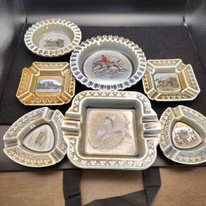 May include: Collection of vintage ashtrays in various shapes, including round, square, and triangular. The ashtrays feature detailed illustrations of landscapes, hunting scenes, and a flying bird, with a color palette of green, brown, and white.