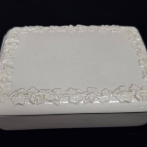 May include: A white ceramic box with a raised floral design around the edge.