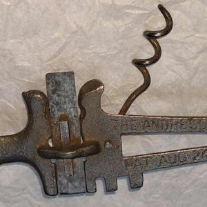 May include: A vintage metal corkscrew with a lever design. The corkscrew has a pointed tip, a handle, and a spiral worm. The text "ANDREWS 1900" and "AUG 24" are visible on the body of the corkscrew. The metal has a tarnished appearance.