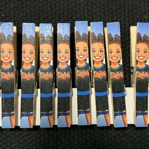 Può includere: Un gruppo di mollette di legno decorate con un design di cheerleader dei cartoni animati. Ogni molletta mostra una cheerleader sorridente in uniforme nera e blu con la scritta "Pride". Lo sfondo è un cielo blu.