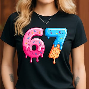 May include: Black t-shirt featuring a colorful graphic of the number 67. The number 6 is pink with sprinkles and dripping icing, while the 7 is blue with sprinkles and an ice cream cone base. The shirt is worn by a person.