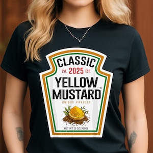 Puede incluir: Camiseta negra con un diseño de etiqueta de mostaza amarilla. La etiqueta dice "CLASSIC YELLOW MUSTARD" y "EST. 2025". Incluye una ilustración de mostaza y el texto "UNIQUE VARIETY". El peso neto es de 368 gramos.