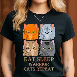 May include: Black t-shirt featuring four cartoon cat faces in orange, gray, brown, and gray. The text "EAT, SLEEP, WARRIOR CATS REPEAT" is printed below the cat illustrations in a colorful font.