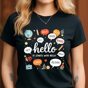 Puede incluir: Camiseta negra con un diseño gráfico colorido. El diseño incluye la palabra "hello" en escritura blanca, rodeada de bocadillos con saludos en varios idiomas, un globo terráqueo y una cámara. También se incluye el texto "IT STARTS WITH HELLO".