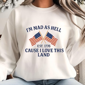 May include: White sweatshirt with the text "I'M MAD AS HELL CAUSE I LOVE THIS LAND" and two crossed American flags. The text "EST. 1776" is also included. The design is in navy blue and red.