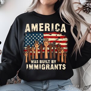 以下が含まれることがあります： 「AMERICA WAS BUILT BY IMMIGRANTS」の白文字がプリントされた黒いスウェットシャツ。さまざまな肌の色の手が前に掲げられたアメリカ国旗のデザイン。スウェットシャツはクルーネックです。