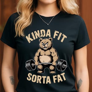 May include: Black t-shirt with a cartoon bear lifting weights. The bear is brown with a stern expression. The text "KINDA FIT" and "SORTA FAT" are printed above and below the bear in a tan color.
