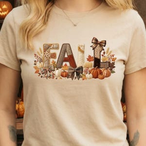 May include: A beige t-shirt featuring the word FALL in large, textured letters with various patterns including plaid, leopard print, and corduroy. The design is decorated with orange pumpkins, brown bows, autumn leaves, wheat stalks, and acorns.