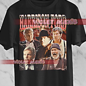 May include: Black t-shirt featuring a vintage-style graphic of Harrison Ford in various roles. The design includes the text "HARRISON FORD" in a retro font. The shirt is hanging on a wooden hanger.