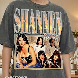 May include: Dark grey comfort colour t-shirt featuring a graphic of Shannen Doherty. The graphic includes the name "Shannen Doherty" in a retro font, and several images of the actress. The text "COMFORT COLOR PEPPER" is printed at the bottom.
