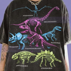 Peut inclure: T-shirt gris foncé avec des squelettes de dinosaures bleus, roses et verts. Les noms des dinosaures sont imprimés : Pteranodon, Brachiosaurus, Triceratops, Tyrannosaurus Rex, Allosaurus, Ankylosaurus et Stegosaurus. Le texte "COMFORT COLORS" et "GRAPHITE" est également visible.