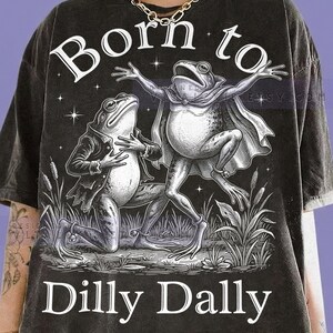 May include: A charcoal gray t-shirt featuring a white graphic illustration of two frogs, one wearing a cape and one in a jacket, surrounded by cattails and stars. The text "Born to" appears above the frogs, and "Dilly Dally" is printed below them.